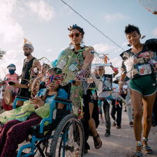 Picture of march through the neighborhood of La Invasion, girl in wheelchair at the front, wearing a butterfly mask, several people wearing costumes in the background. Photo by Ema Fantini